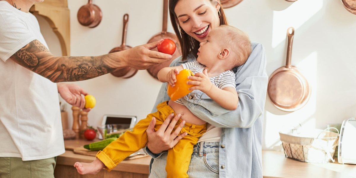 Two adults in a kitchen holding a toddler who is reaching for fruit