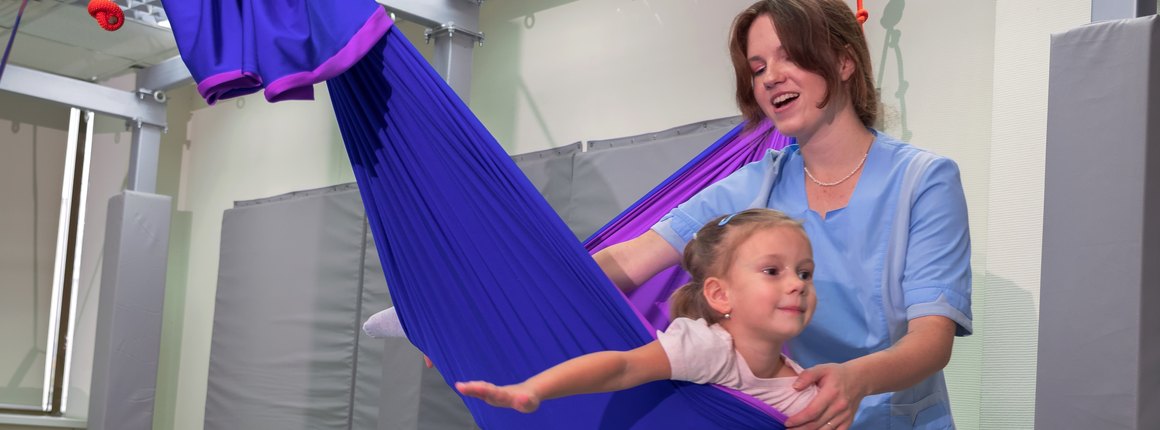 A young child lying in a therapy swing while a therapist supports the movement
