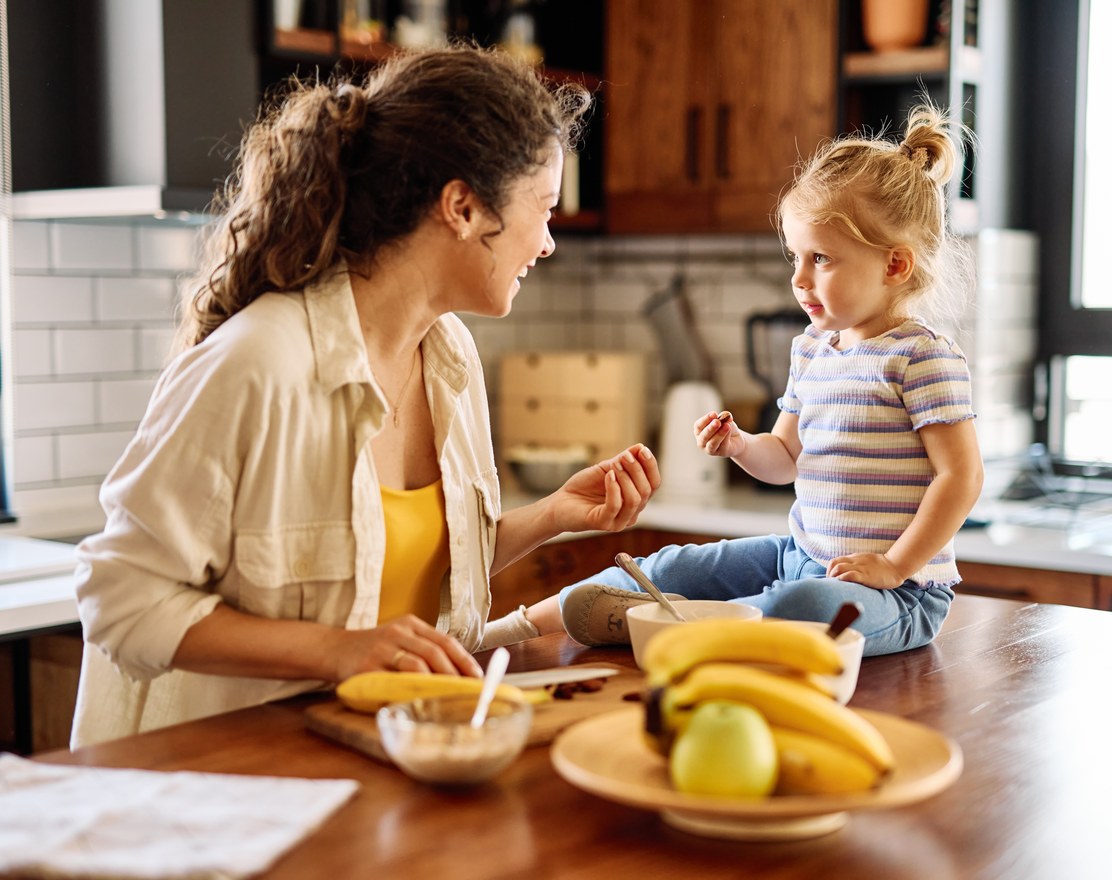 A mother and toddler in a kitchen with a bowl of fruit on the table