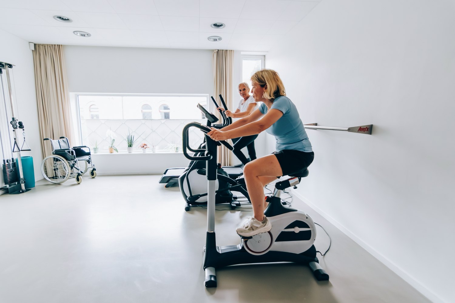Two people exercising on stationary bikes in a bright rehabilitation gym, supervised by a therapist.
