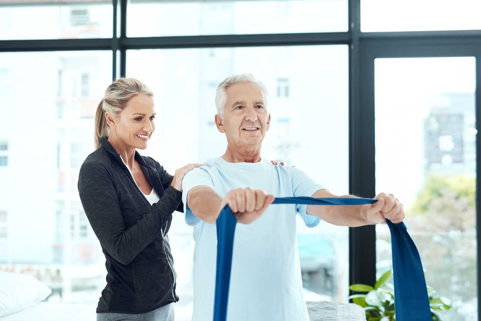 A physical therapist supporting an older adult through a resistance band strengthening exercise in a bright therapy room.