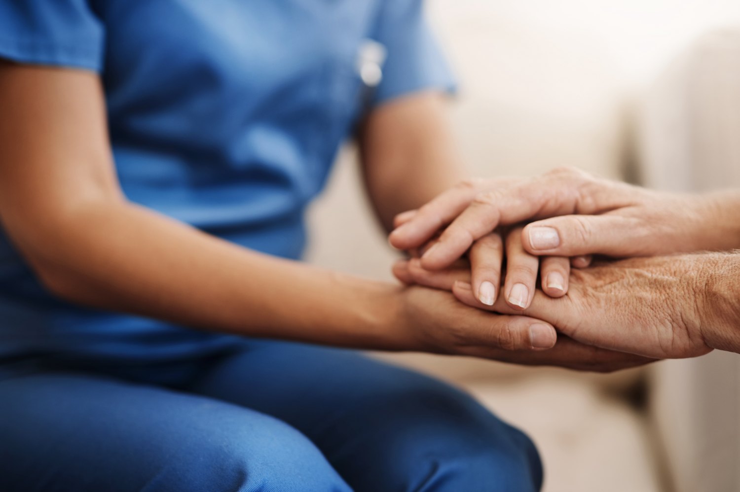 A clinician in scrubs holding an older patient's hands during an unhurried conversation.