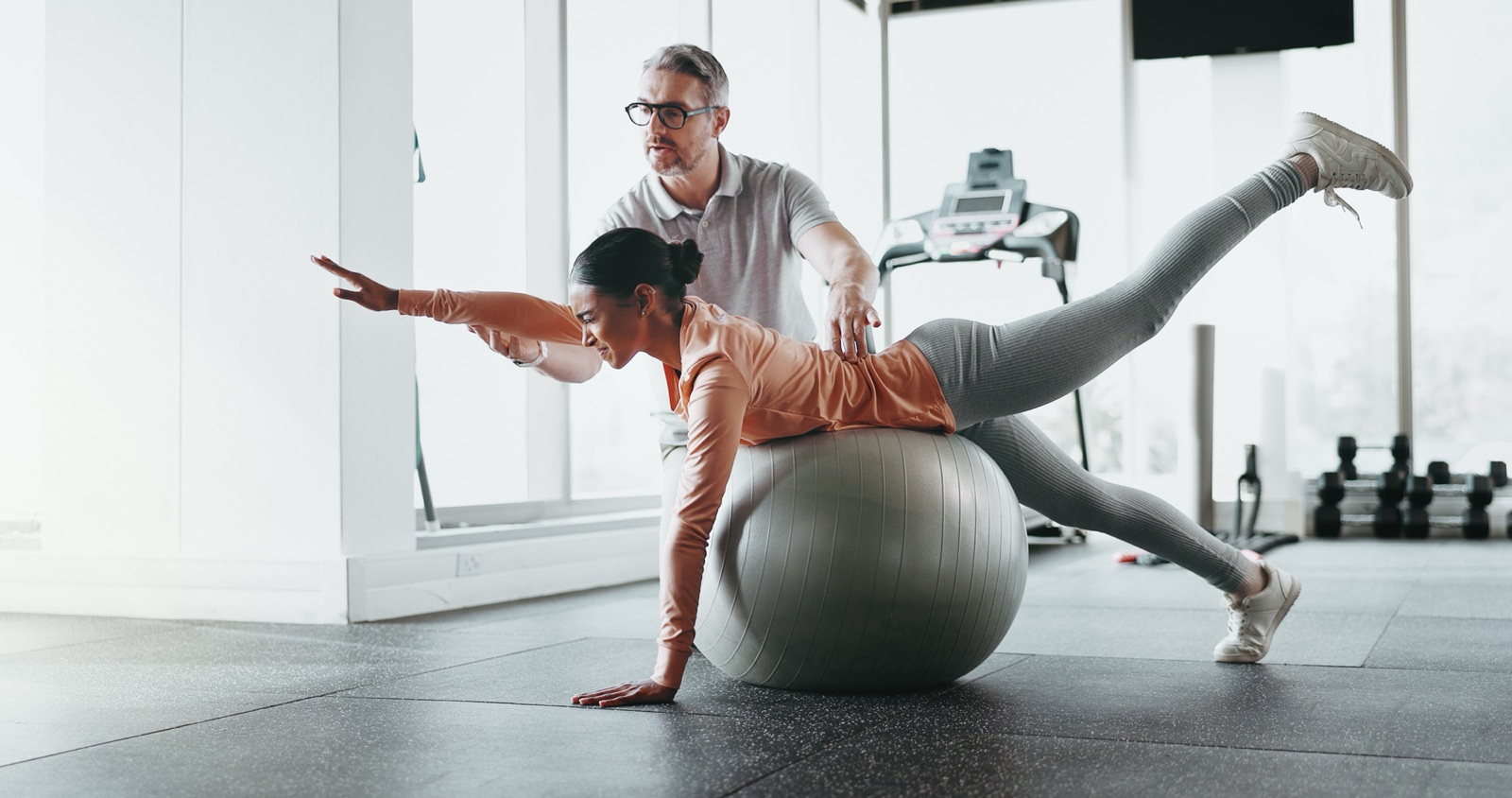 A physical therapist supervising an adult practicing balance and core control over an exercise ball in a rehabilitation gym.