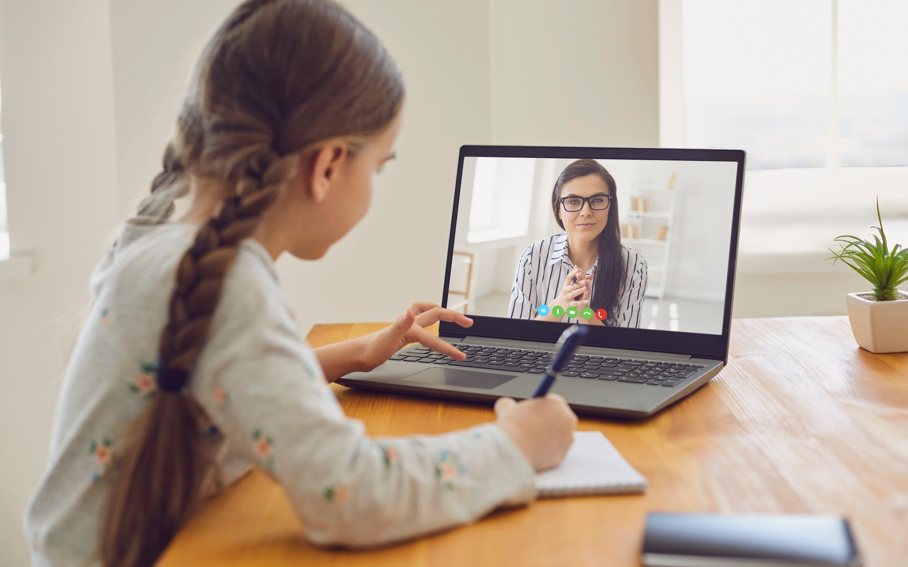 A girl at a desk taking notes during a video call with her teacher on a laptop