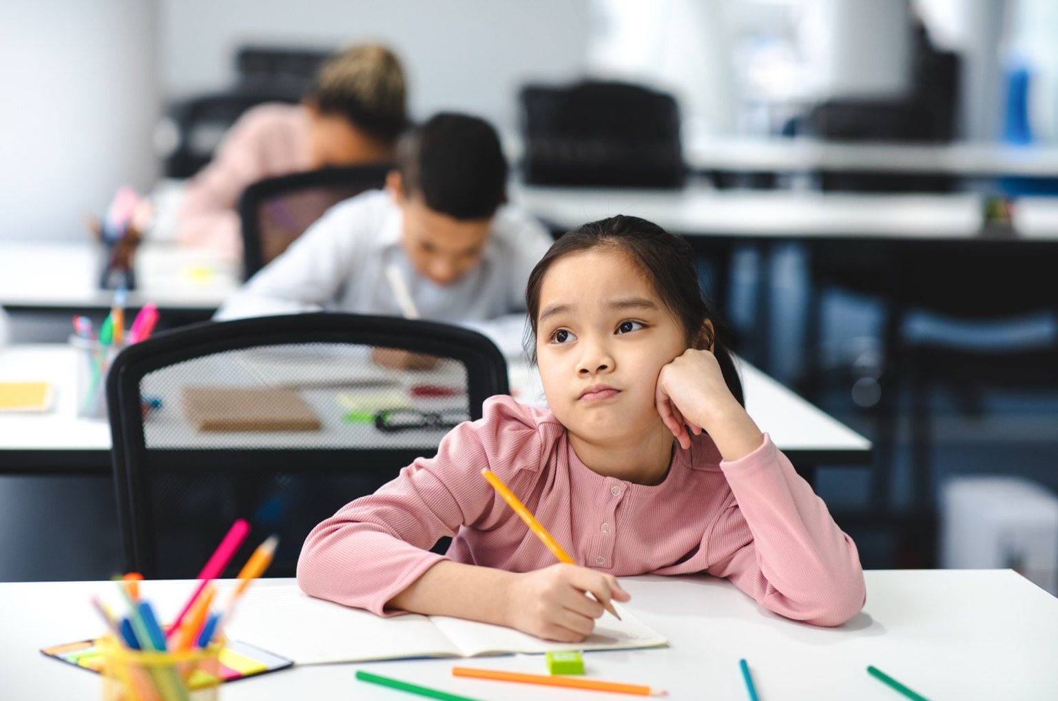 A girl at a classroom desk gazing away while the children behind her carry on working