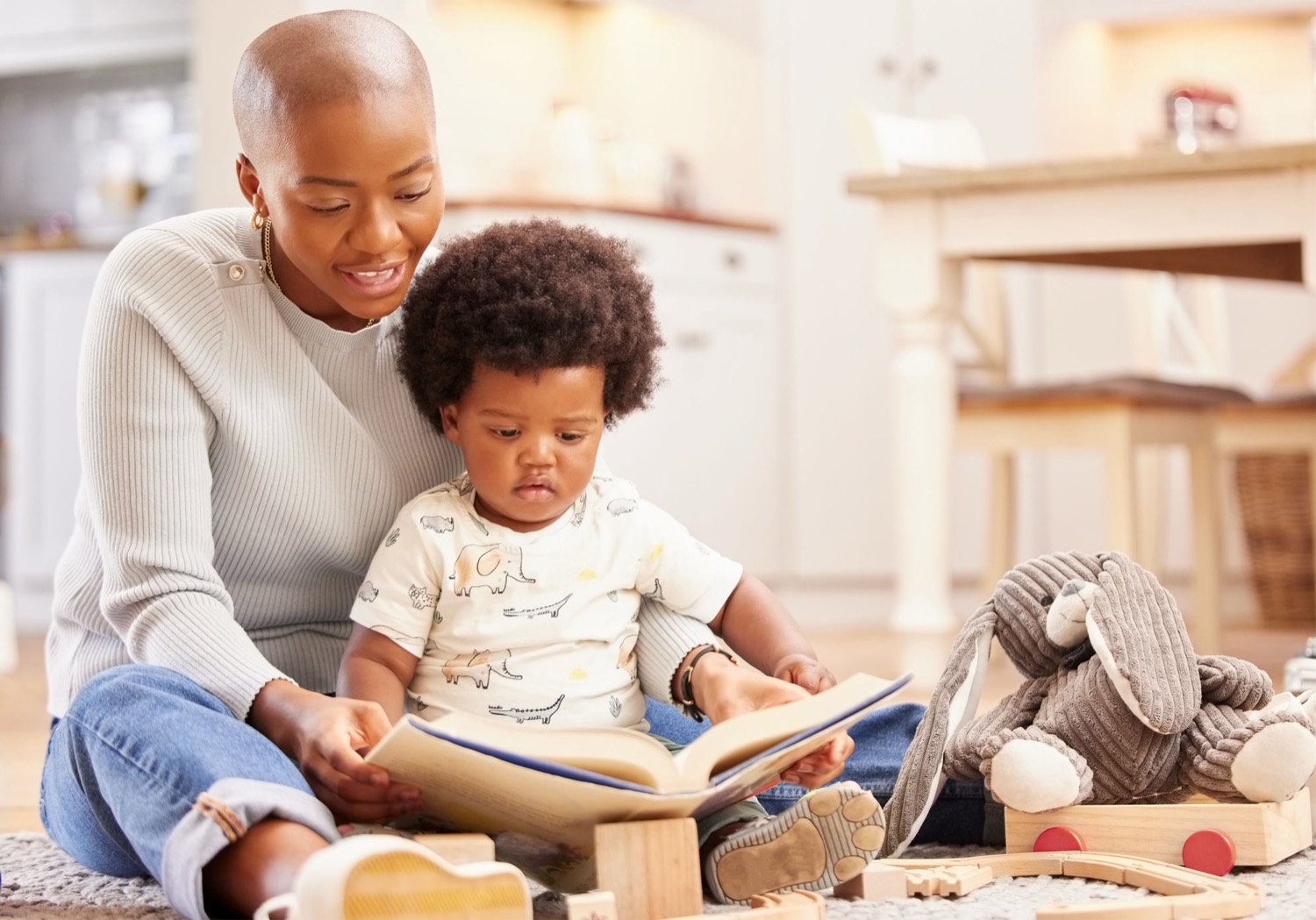 A mother and her baby sitting on the floor together, looking at a picture book surrounded by wooden toys