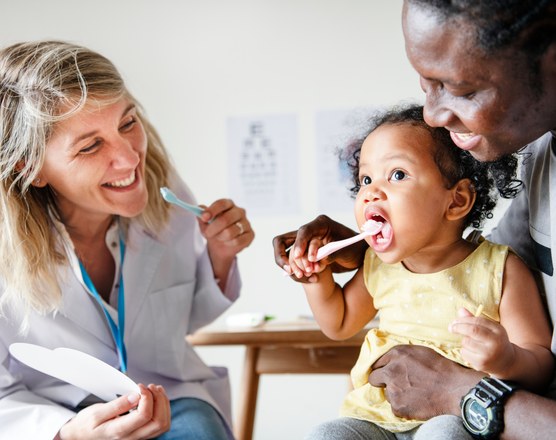 A dentist showing a toddler how to brush their teeth while a parent holds them
