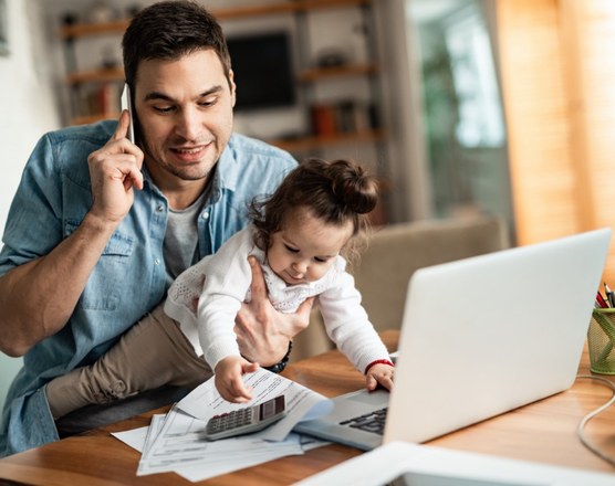 A father working at a laptop at home with his toddler beside him