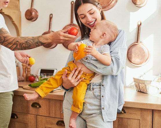 Two adults in a kitchen holding a toddler who is reaching for fruit