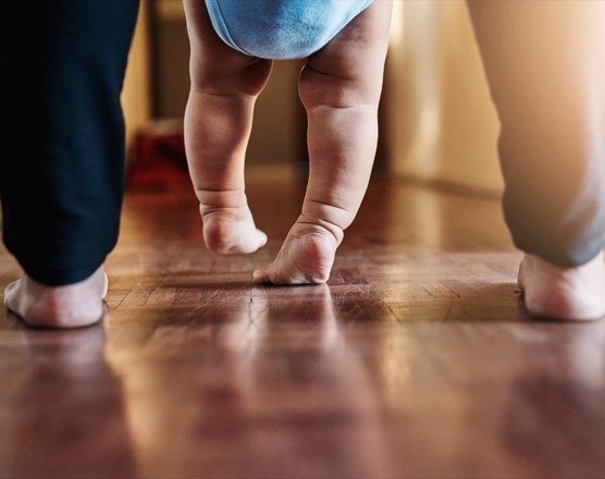 A baby's bare feet taking steps on a wooden floor, held by a parent