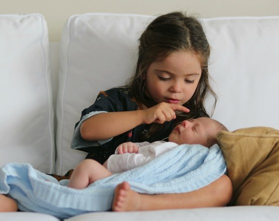 An older sister sitting with a sleeping swaddled newborn across her lap, raising a finger to her lips