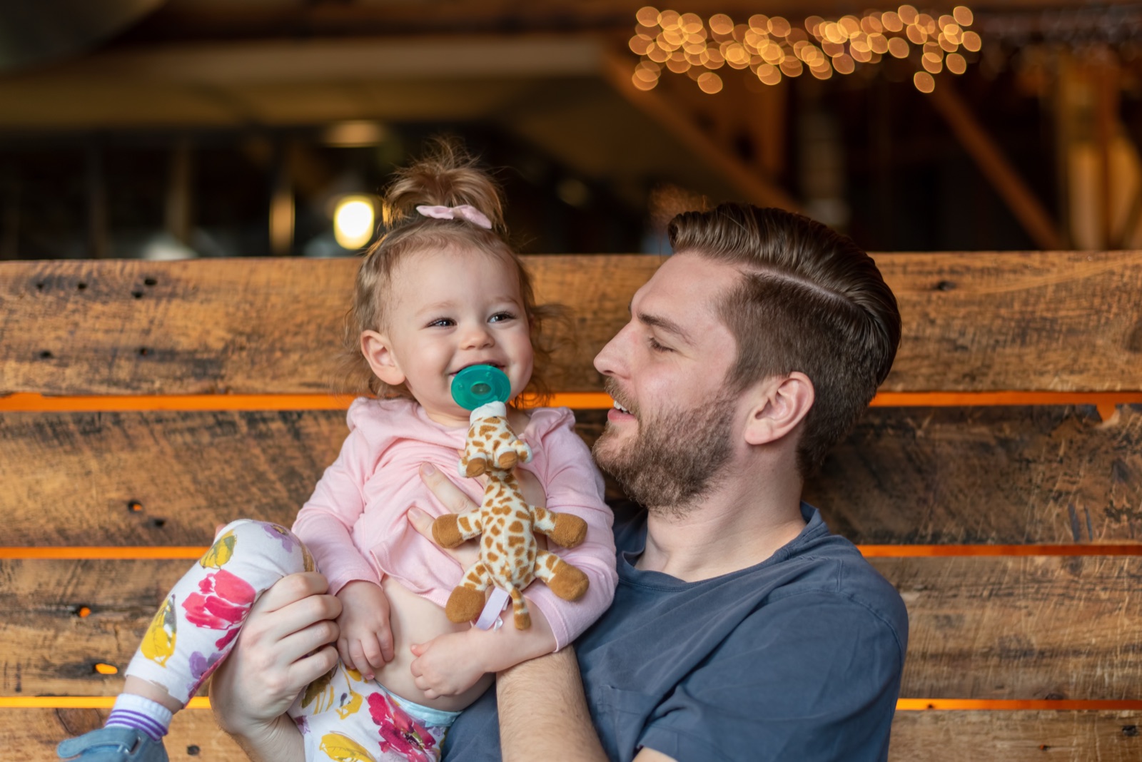 A father holding a smiling toddler who has a pacifier and a soft giraffe toy