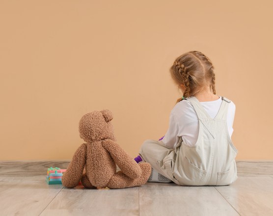 A young girl sitting on the floor with her back turned, beside a teddy bear