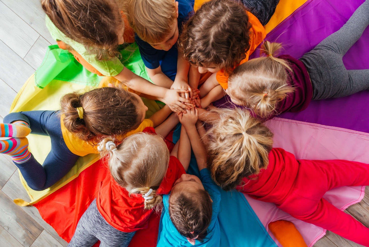 Young children sitting in a circle on a colored play parachute with their hands joined in the middle