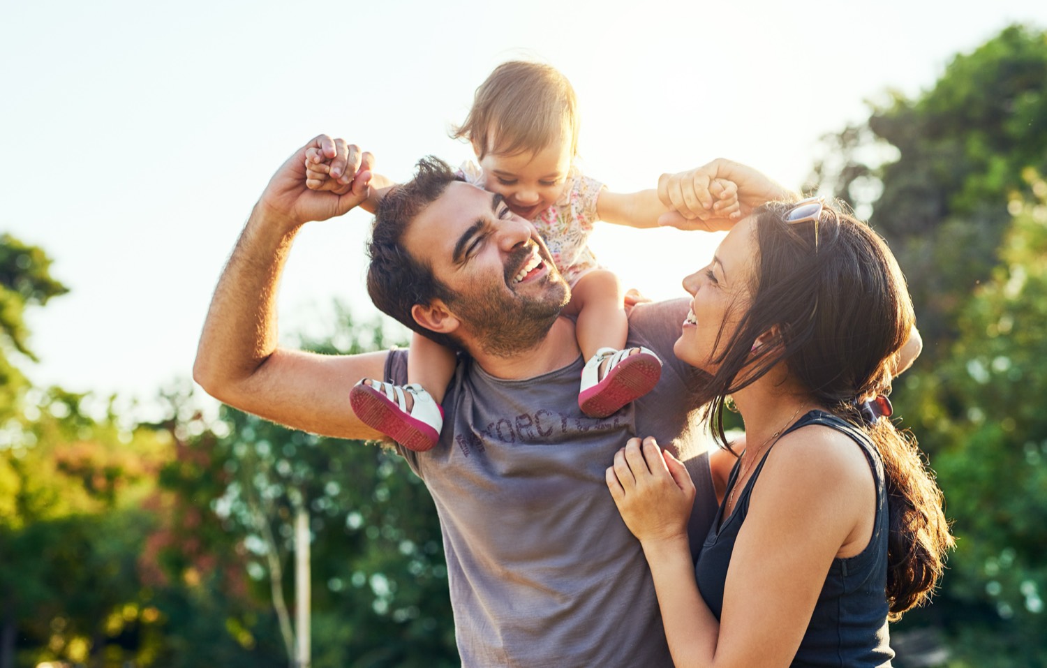 A father carrying his laughing toddler on his shoulders with her mother beside them outdoors