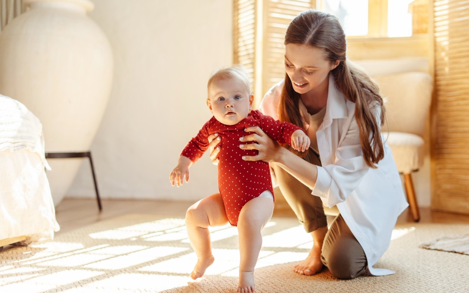 A mother kneeling on a rug holding a baby under the arms as the baby takes steps toward the camera
