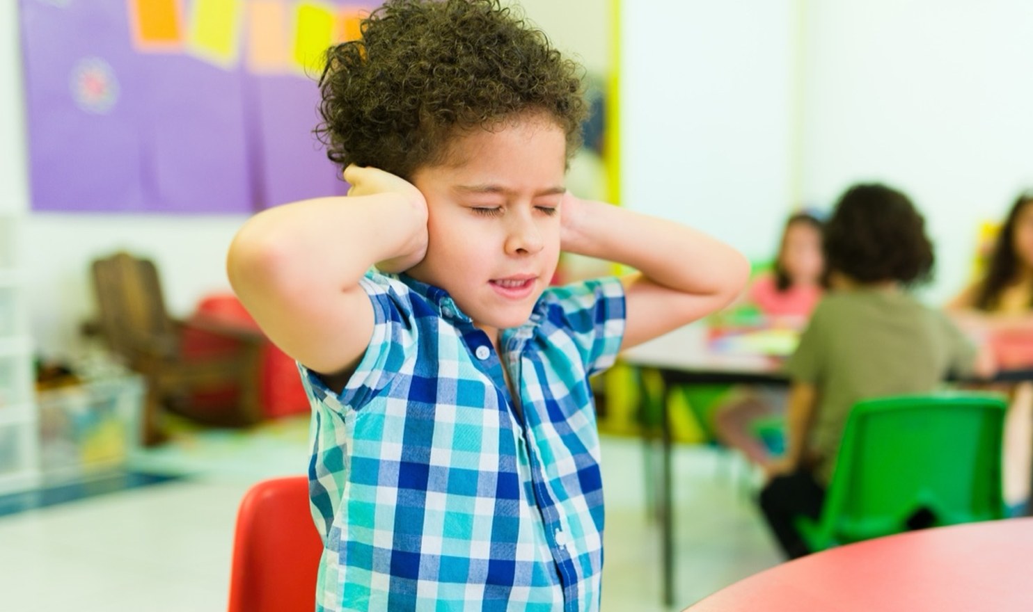 A boy at a classroom table covering both ears while other children work behind him