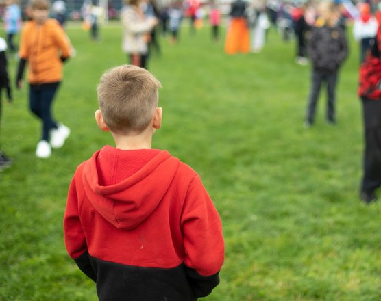 A boy in a red hooded top standing apart from a crowd of children running in a park