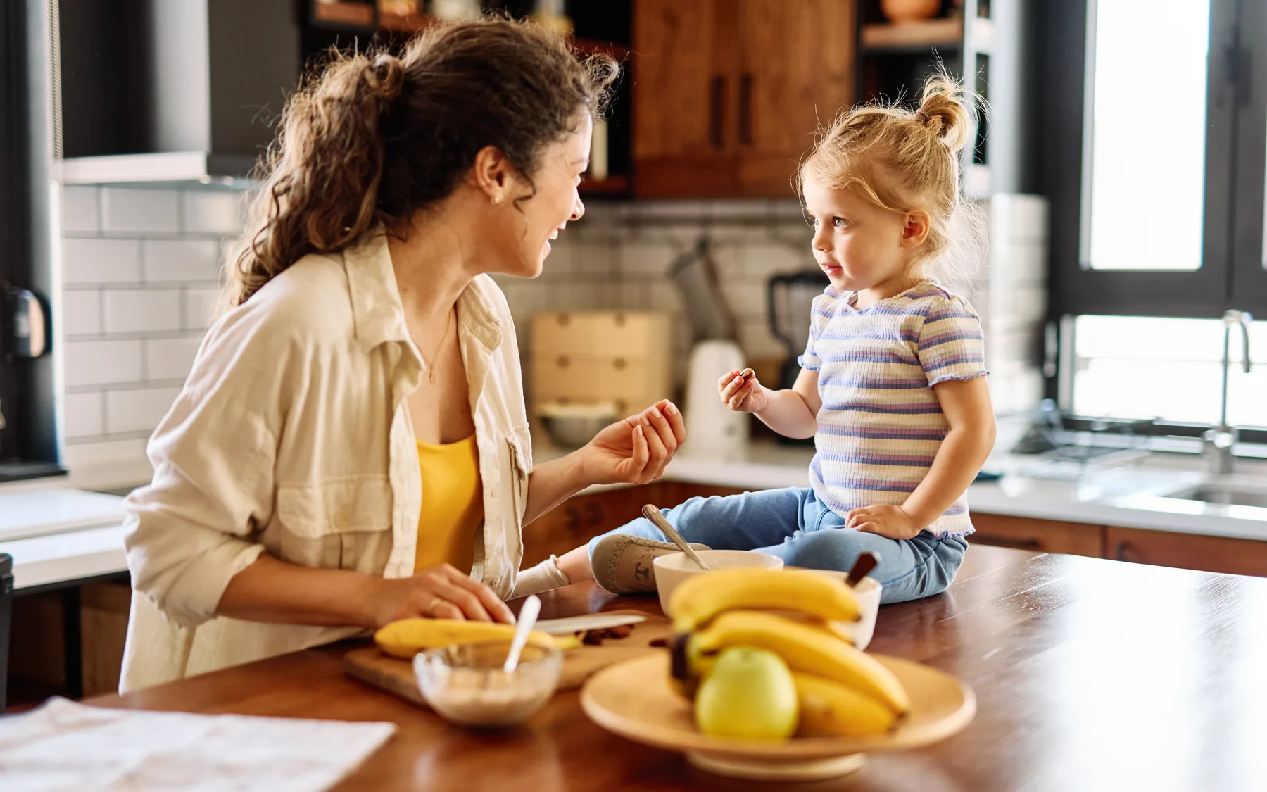 A toddler sitting on a kitchen counter eating finger food while a parent talks with her