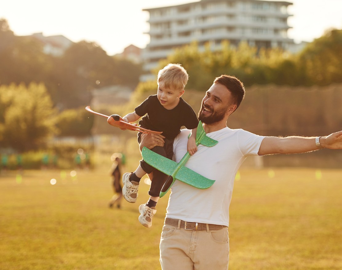 A father carrying his son through a park, the boy holding a toy plane