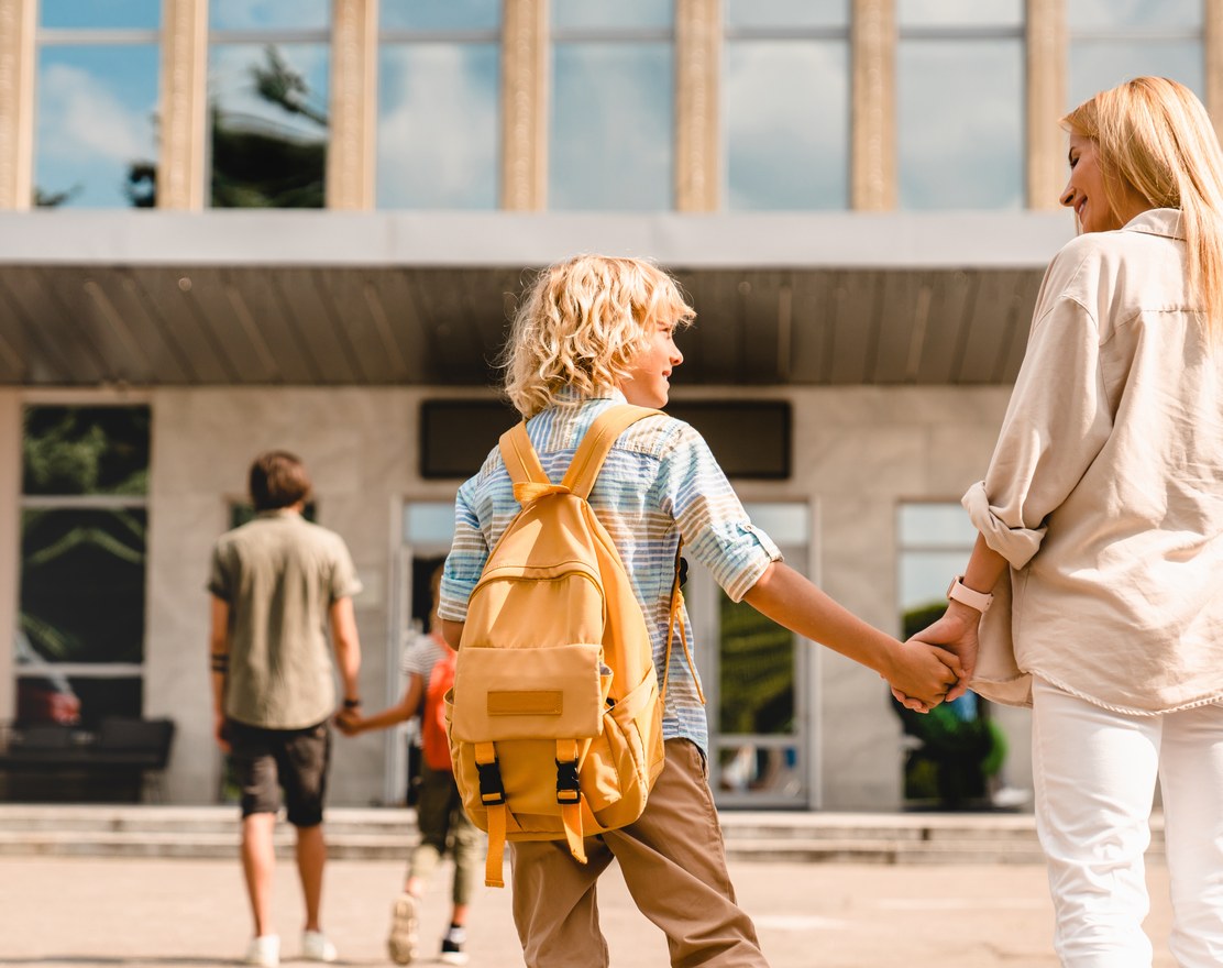 A parent and child walking toward a school building, the child wearing a backpack