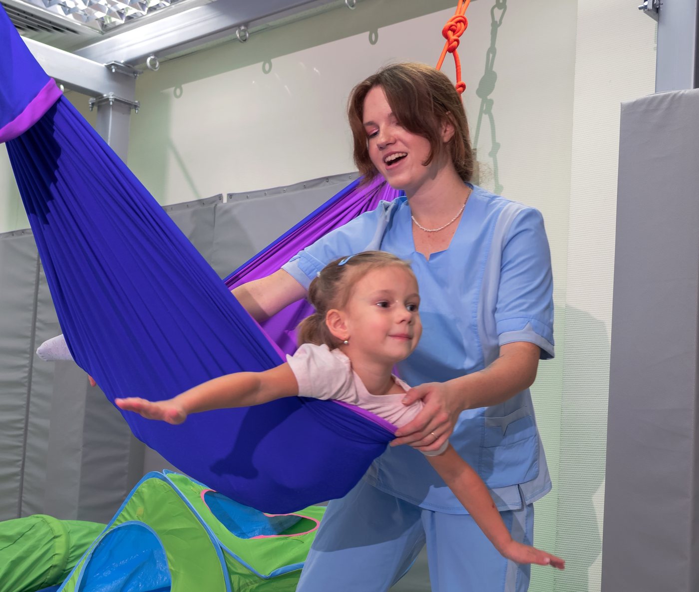 An occupational therapist supporting a young girl in a therapy swing in a sensory gym