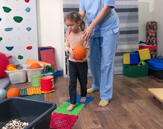 A preschool-aged girl stepping across textured balance tiles while a physical therapist steadies her shoulders in a therapy gym