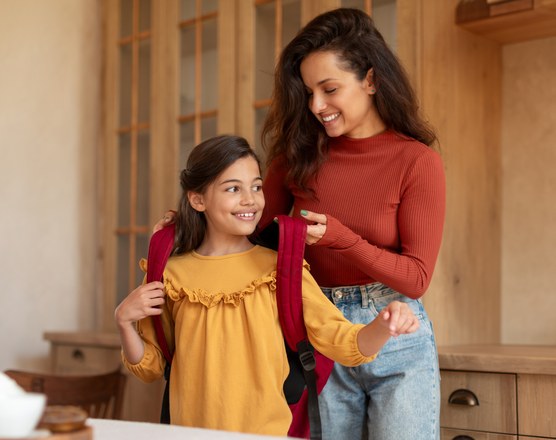 A mother helping her daughter into a school backpack in the kitchen