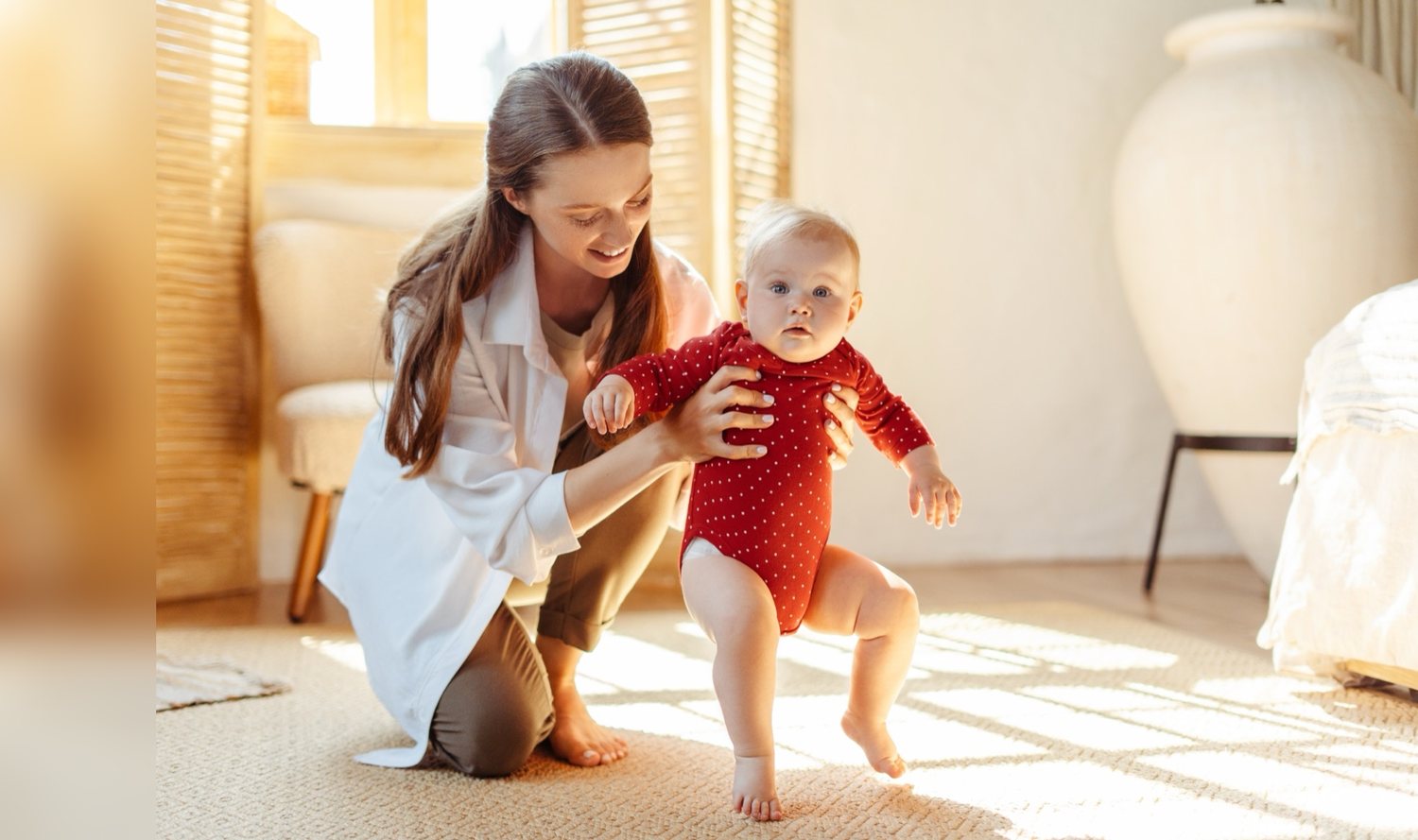 A mother crouching behind her baby, supporting the baby’s chest as the baby takes steps