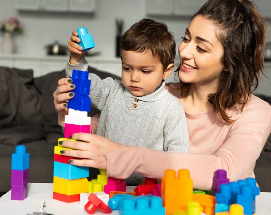 A mother watching a toddler stack coloured building blocks into a tower