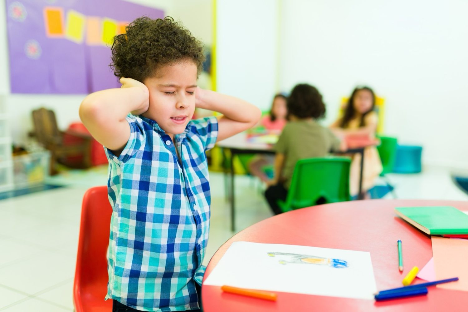 A boy at a classroom table covering both ears with his eyes shut while other children work behind him