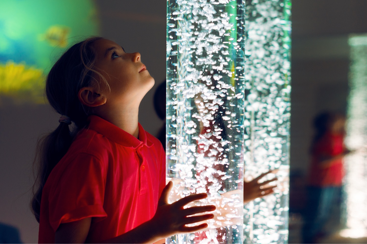 A child watching bubbles rise through a lit sensory tube in a darkened room