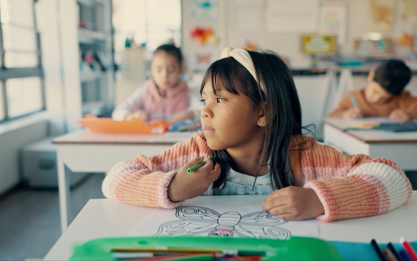 A girl at a classroom desk looking away from her work while other children keep working behind her