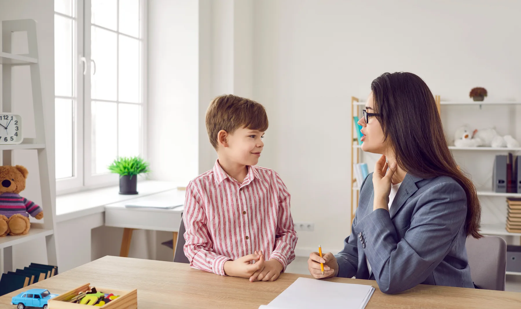 A young boy sitting across a table from a clinician who is taking notes during an assessment
