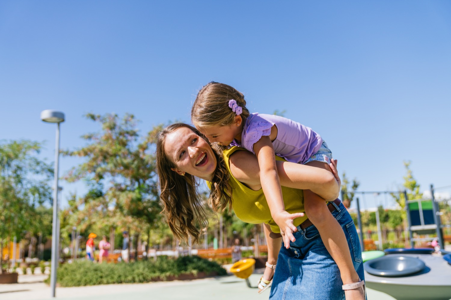 A mother giving her daughter a piggyback at a playground on a sunny day