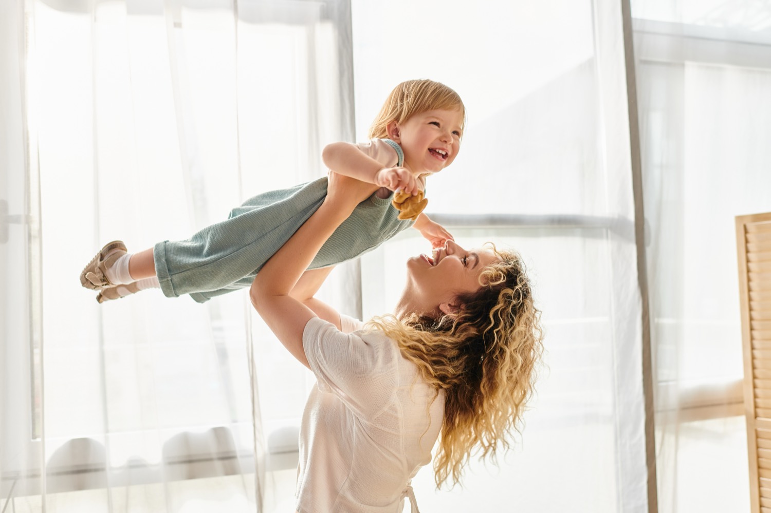 A mother lifting her laughing toddler into the air in a bright room