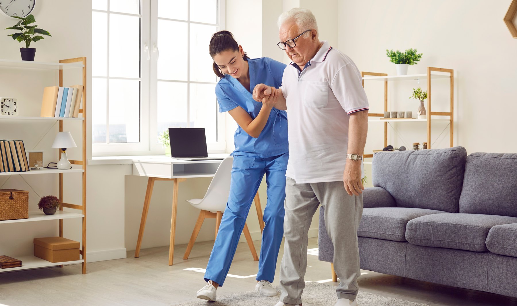 A physical therapist supporting an older man by the arm as he walks across a room