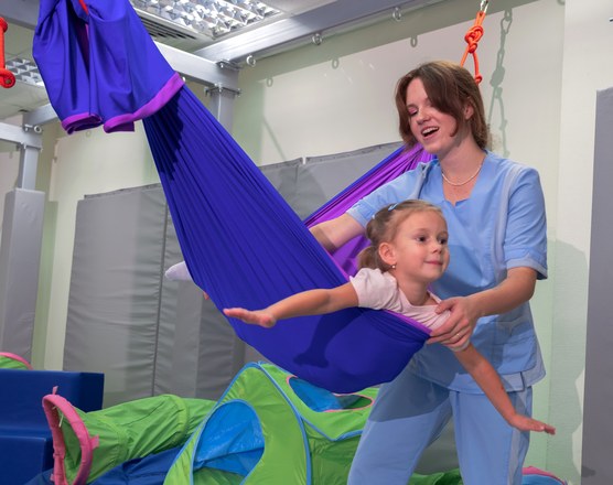 A child lying in a therapy swing while a therapist guides the movement