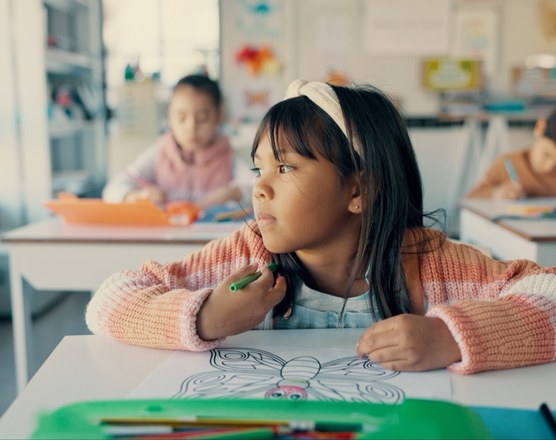 A girl in glasses looking up from a drawing at a classroom desk