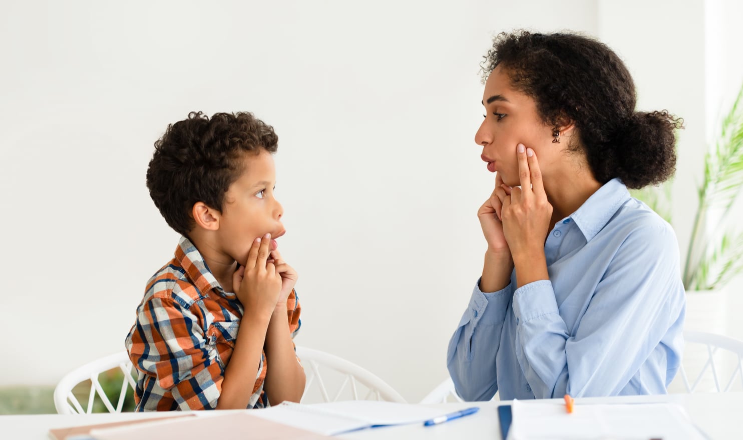 A speech-language pathologist and a school-age boy sitting face to face at a table, both shaping a sound with their hands at their mouths