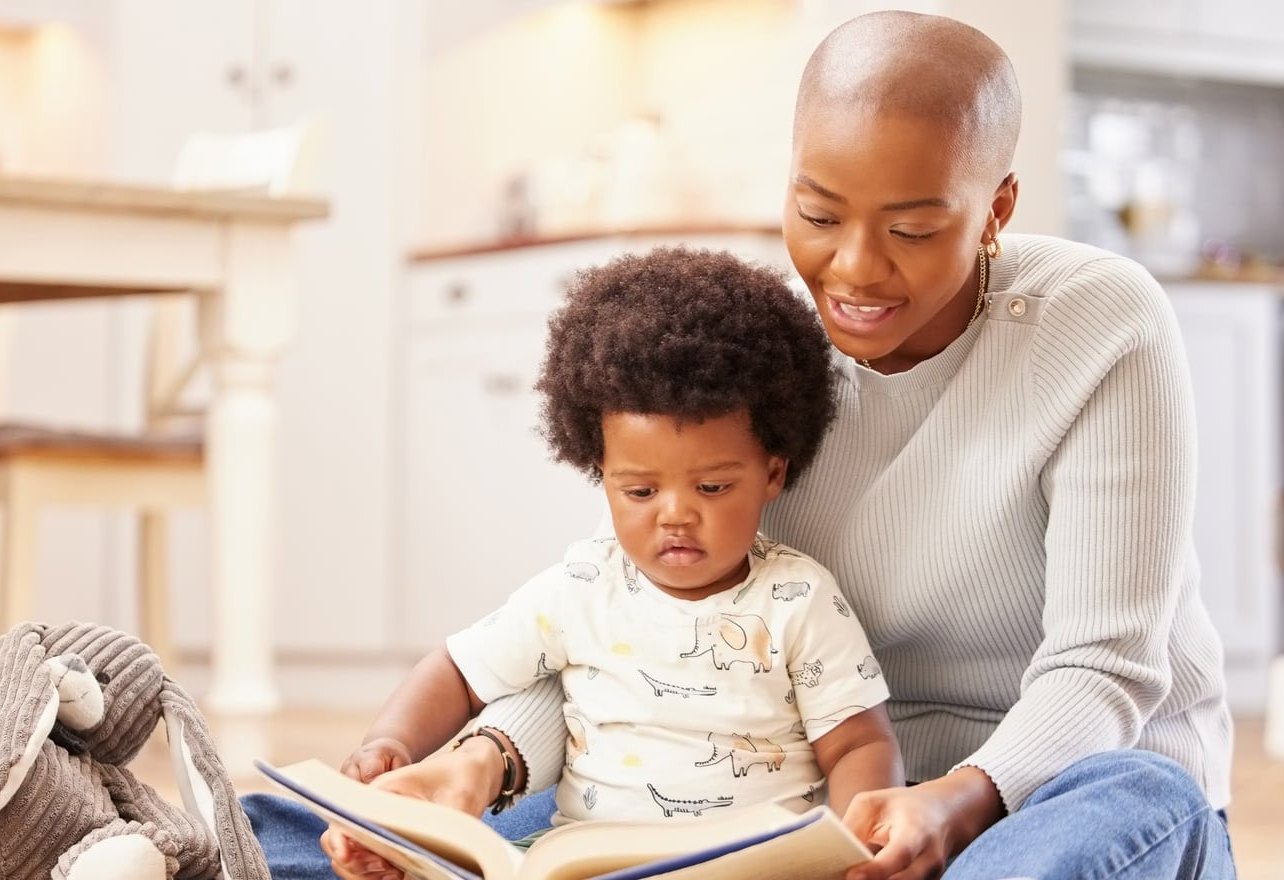 A parent sitting on the floor with a toddler in her lap and an open picture book across their knees