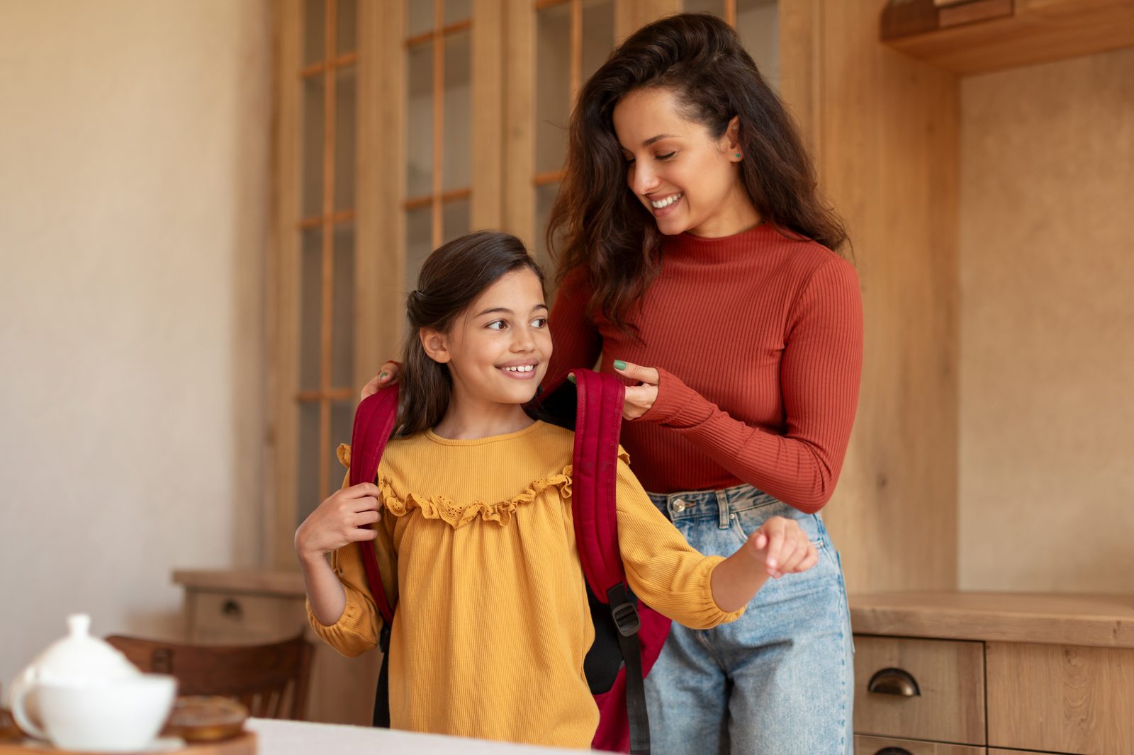 A smiling mother helping her daughter put on a red school backpack in the kitchen