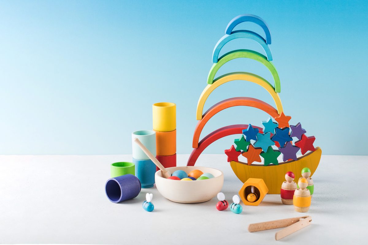 Wooden toys in rainbow colors: a stacking rainbow, a balancing boat of stars, nesting cups, a bowl of balls and peg figures
