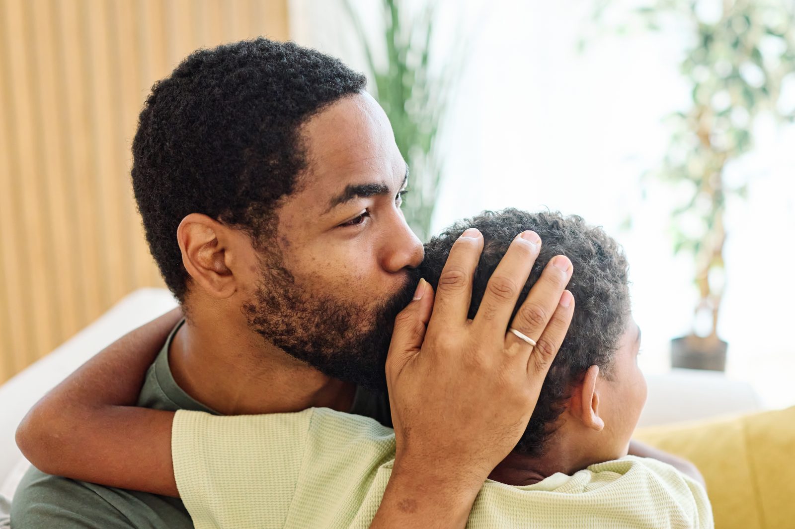 A father hugging his young son and kissing the top of his head, one hand cupped over the boy's ear
