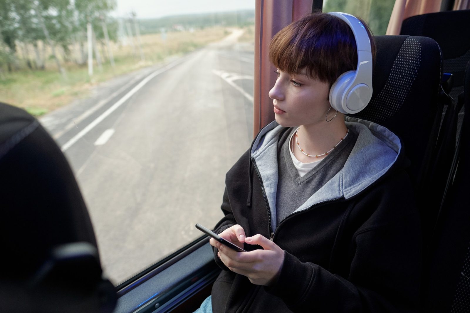 A teenager wearing large white headphones looking out of a bus window, holding a phone