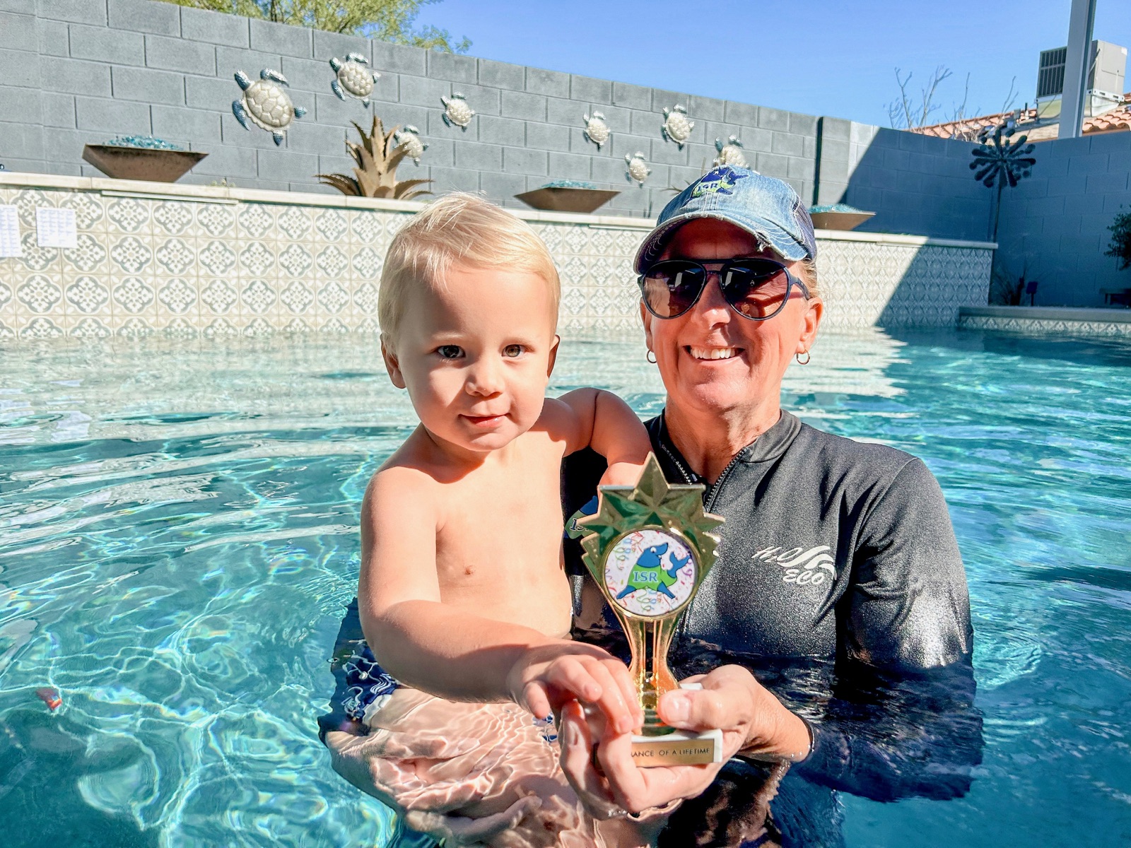 A toddler held by a swim instructor in a pool, holding a small trophy