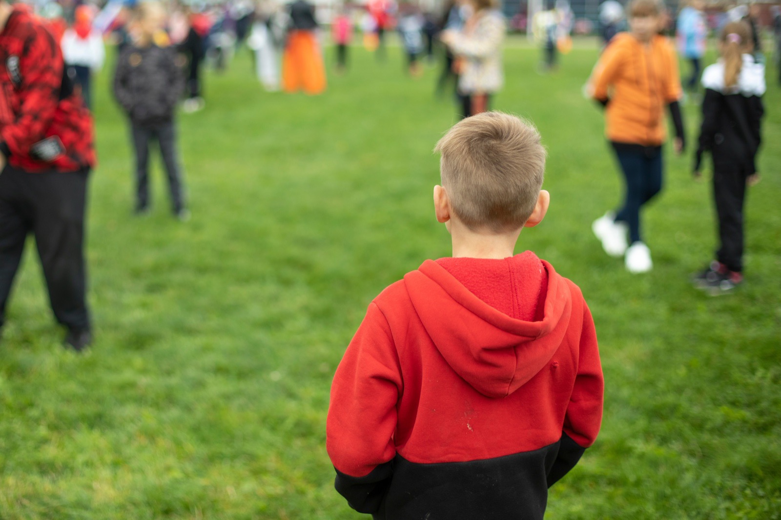 A child in a red hooded top standing apart from a group of other children on a school field, seen from behind.