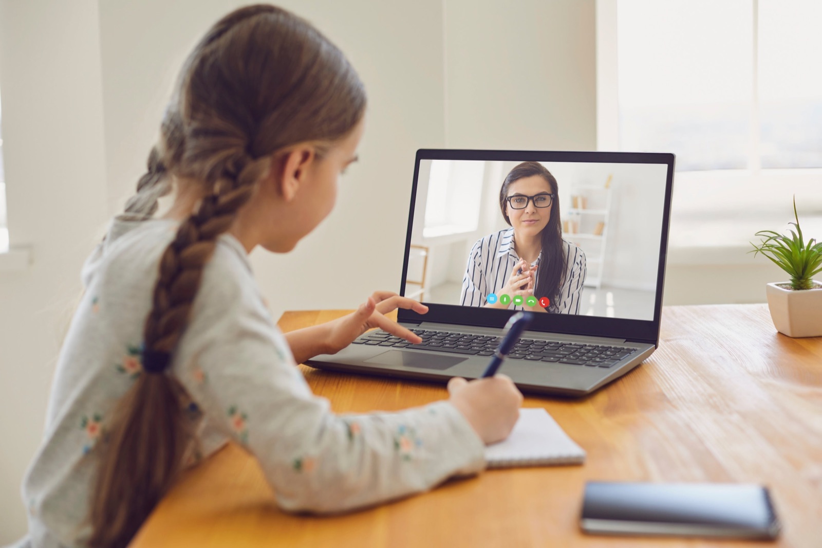 A girl at a wooden table writing in a notebook while a speech therapist speaks to her from a laptop screen