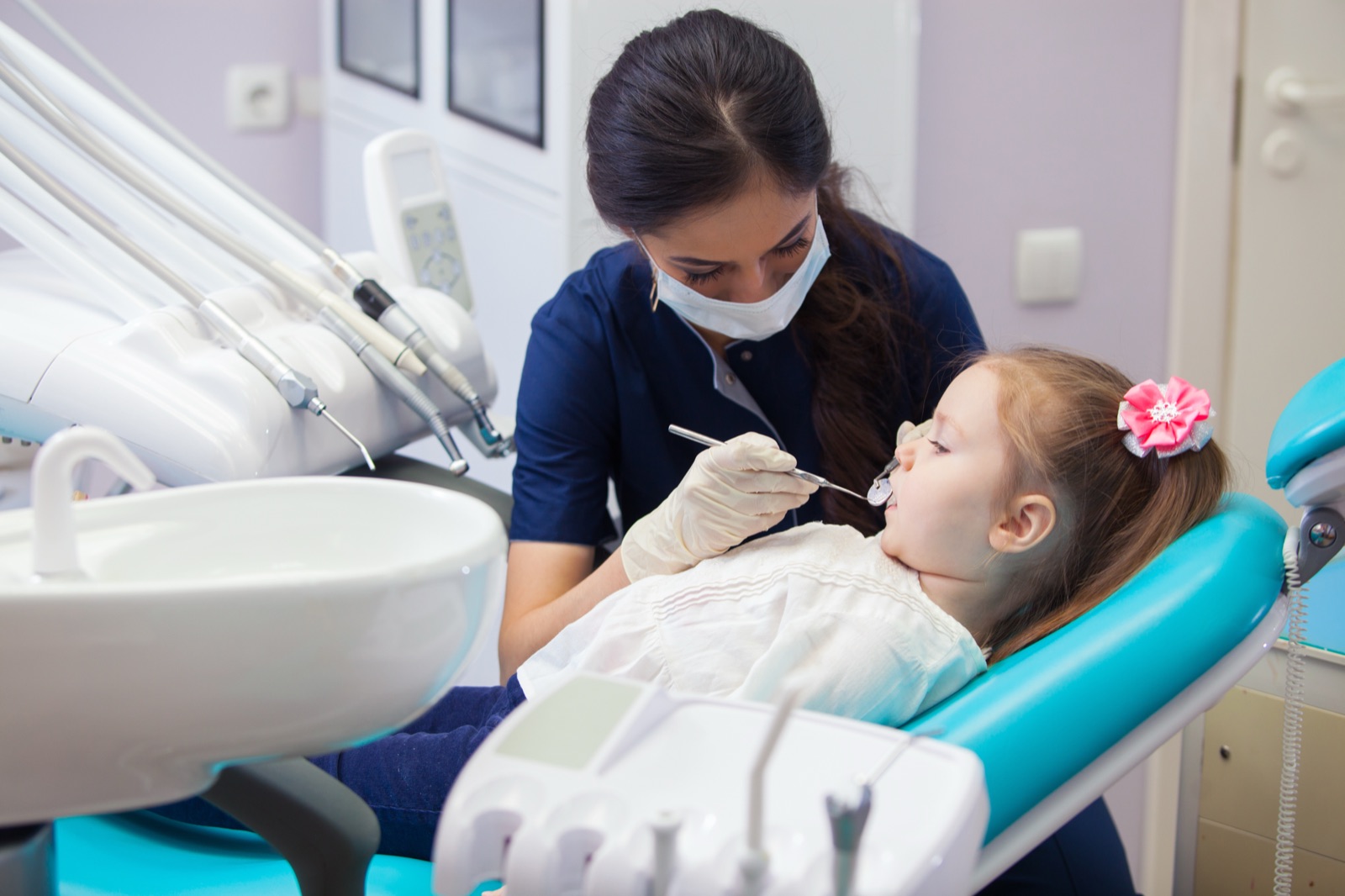 A dentist in a mask examining a young girl's front teeth with a dental mirror while she lies back in the chair