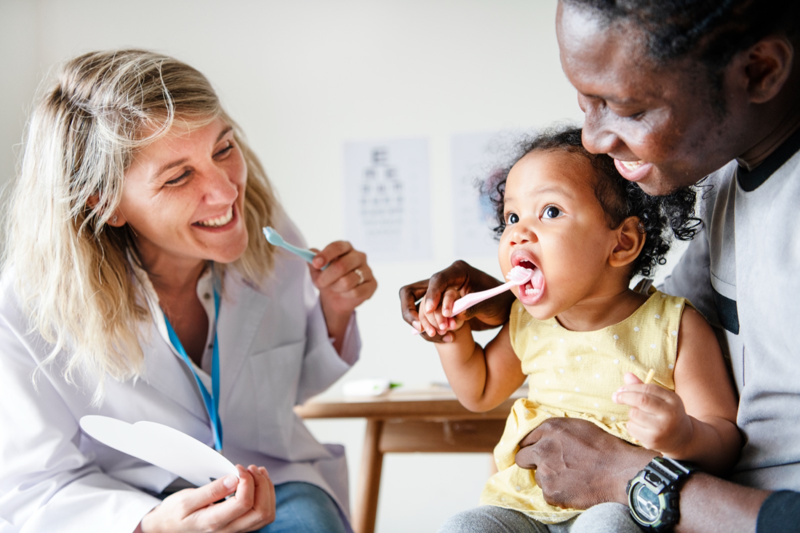 A dentist and a father showing a toddler on her father's lap how to brush, the child holding a pink toothbrush to her own teeth